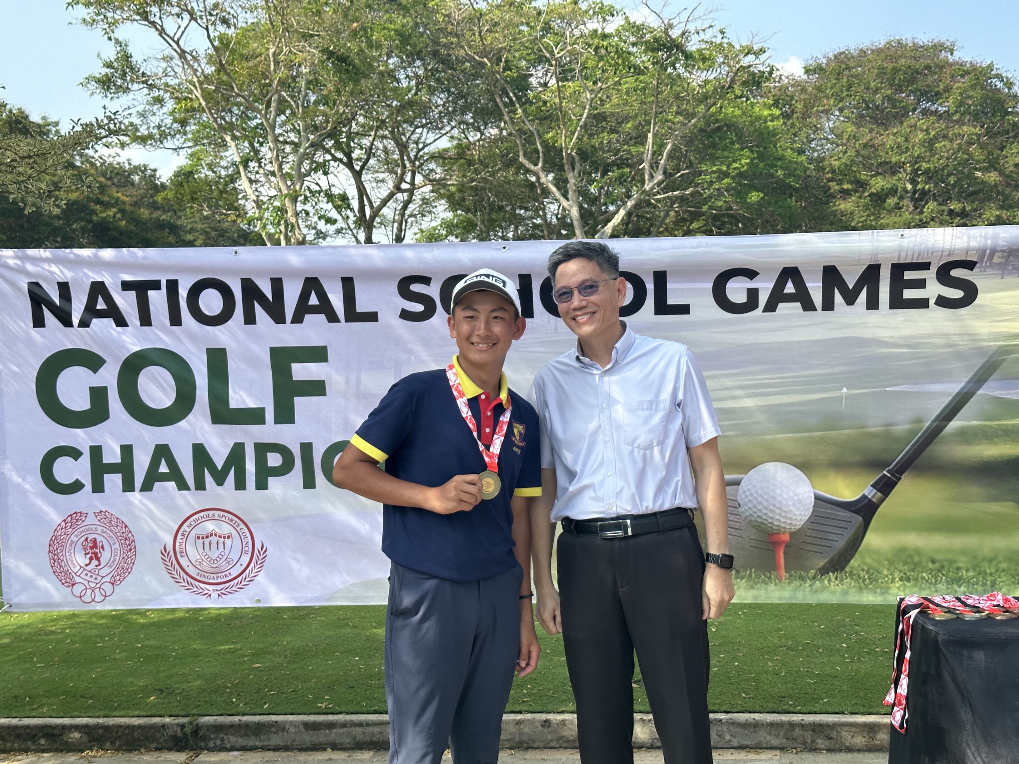A young boy holding a gold medal poses with an adult man in front of a National School Games Golf Championship banner.